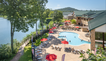 A pool with red umbrellas, lounge chairs, and people, next to a building by a lake, surrounded by greenery and a scenic view.