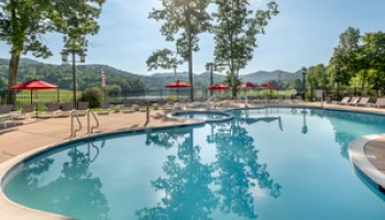 A serene outdoor pool is surrounded by lounge chairs, trees, and red umbrellas, set against a backdrop of mountains and a clear blue sky.