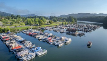 A marina with numerous docked boats on calm water, surrounded by lush greenery and hills under a clear blue sky.