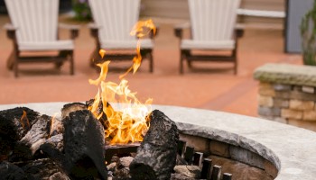 A cozy outdoor fire pit with burning logs and three Adirondack chairs in the background, set on a patio.