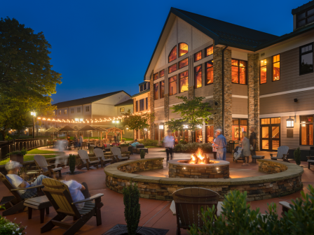 A warm outdoor gathering spot with a fire pit, Adirondack chairs, and a well-lit building in the evening.