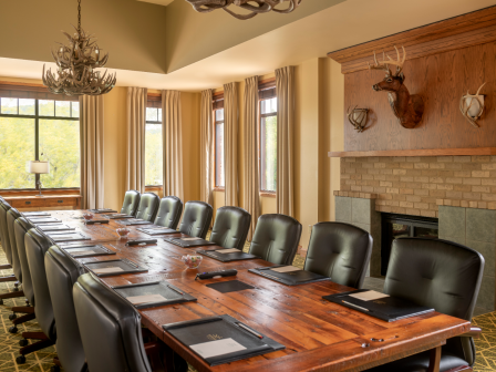 A conference room with a long wooden table, leather chairs, large windows, antler chandelier, and a fireplace with deer decor on the wall.