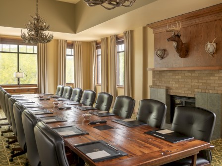 A spacious conference room features a long wooden table, black chairs, a fireplace with a mounted deer head, and large windows with curtains.