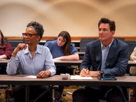 A group of people are seated in a classroom setting, engaged and taking notes during what appears to be a meeting or presentation.